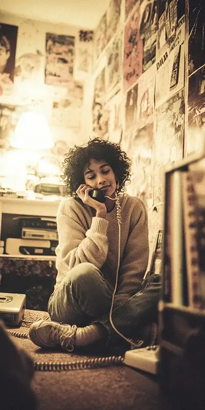 a young woman sitting on the floor of her bedroom talking on a corded landline telephone